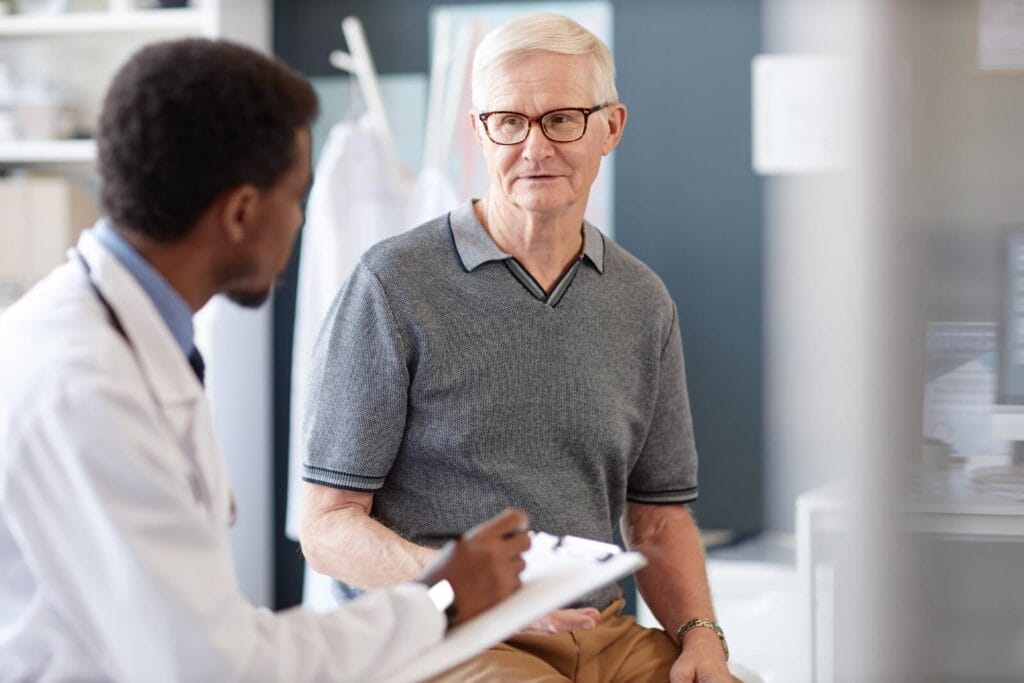 An older adult man talks with a doctor, who is writing on a clipboard.