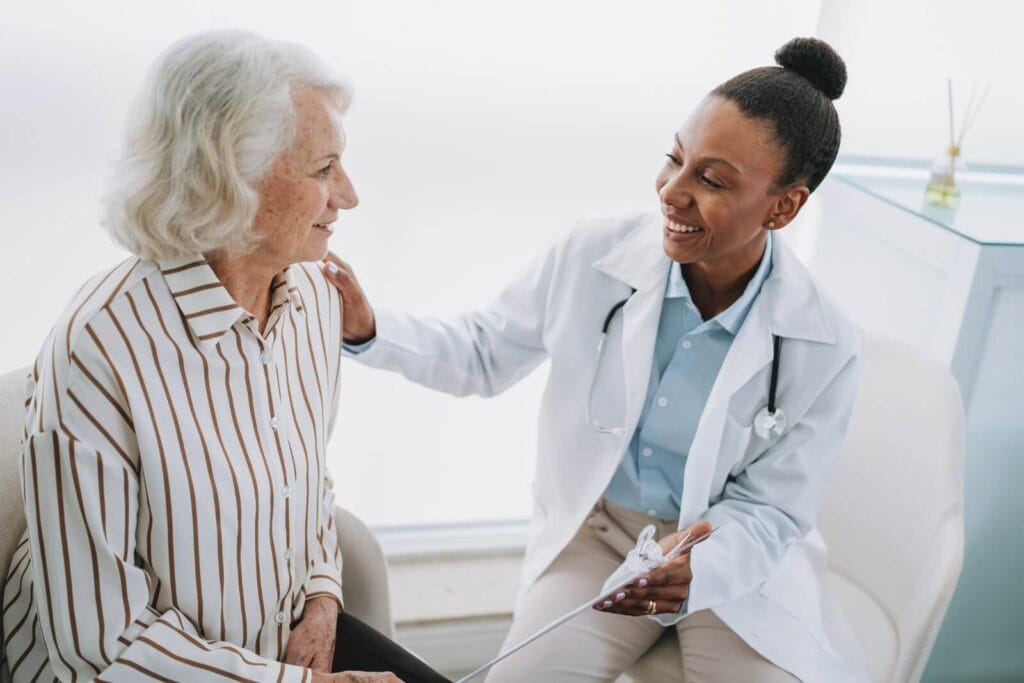 An older adult woman sits with a woman doctor who is holding a clipboard and placing her hand on the other woman's shoulder. They are smiling at each other.
