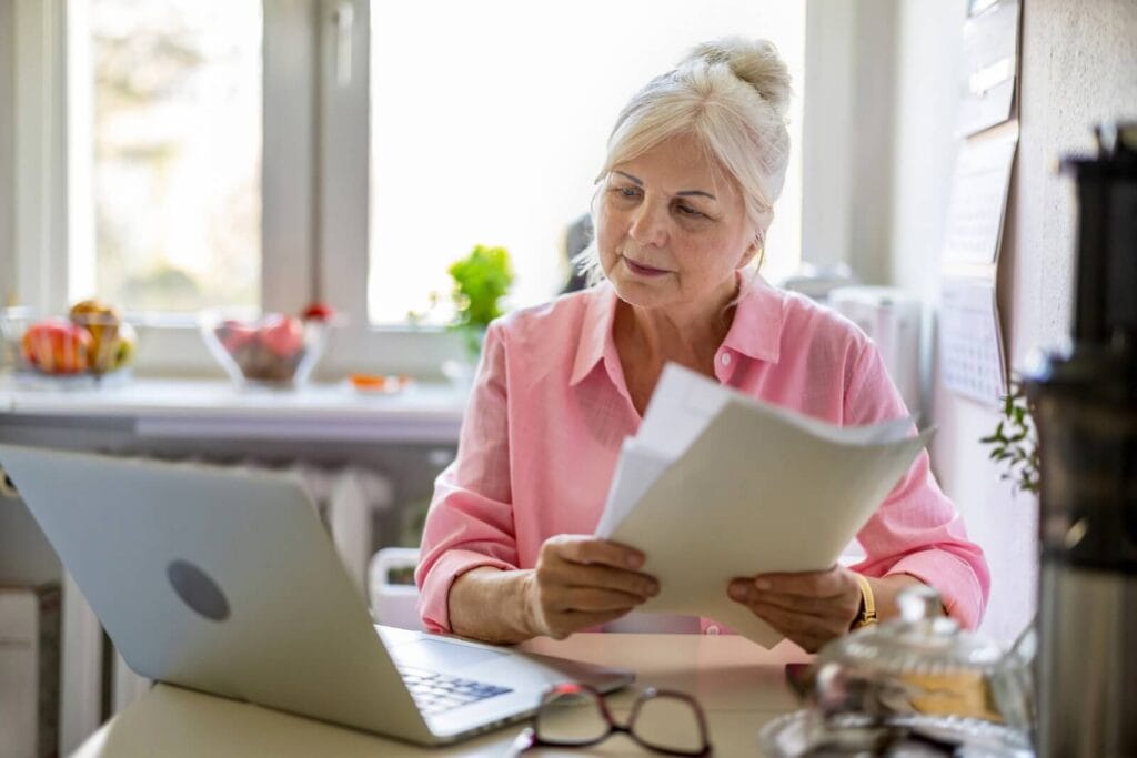 An older adult woman sits at a table with a laptop on it, holding some papers.