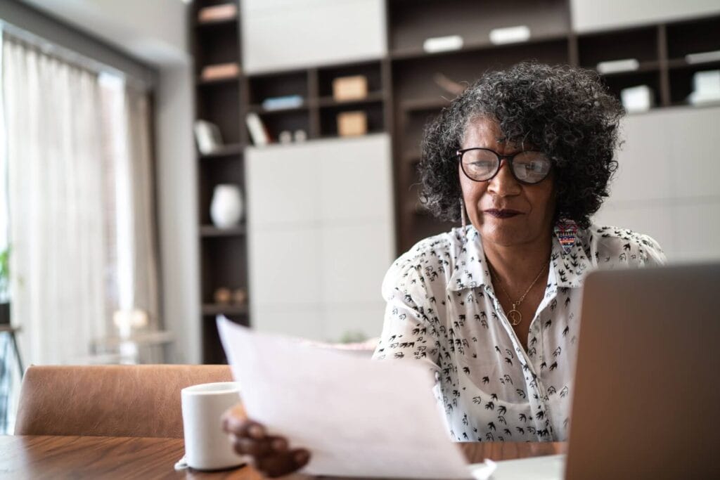An older adult woman sits at a table with a laptop in front of her. She is holding and looking at a piece of paper.