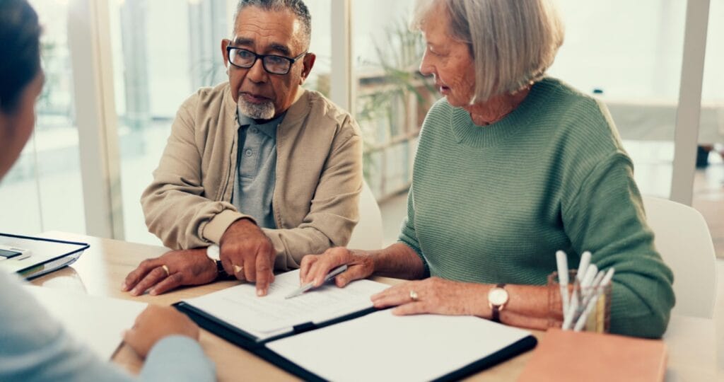 An older adult man and woman sit across a desk from a lawyer. They have a binder with paperwork in front of them.