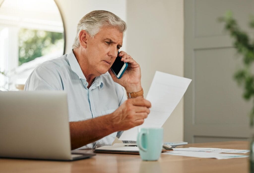 An older adult man is sitting at a table, speaking on the phone, and looking at a piece of paper. He looks serious. There are also a laptop, more paperwork, and a coffee mug in front of him.