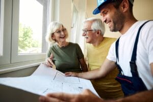An older adult man and woman stand next to a contractor, who is showing them a floor plan for modifying their home. Everyone is smiling.
