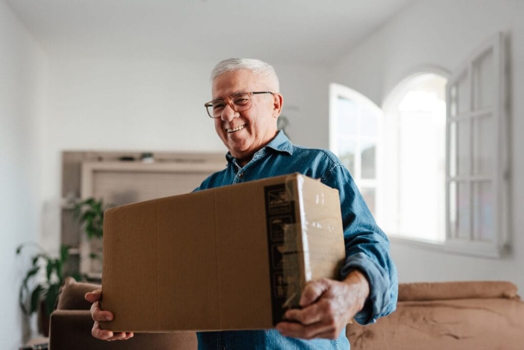A smiling older adult man holds a cardboard box. He is smiling.