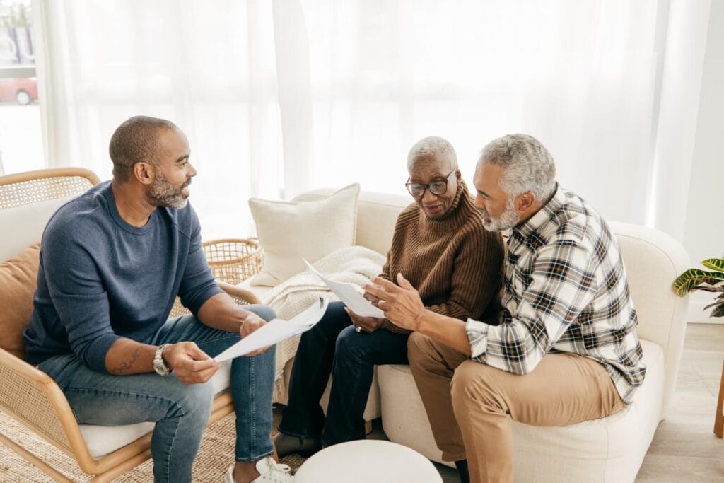 An older adult man and woman are sitting in a living room with a younger man, looking over paperwork,