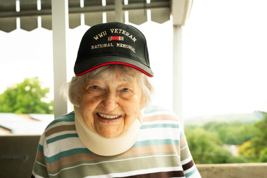 An older adult woman is smiling at the camera. She is wearing a hat that reads "WWII VETERAN, NATIONAL MEMORIAL."