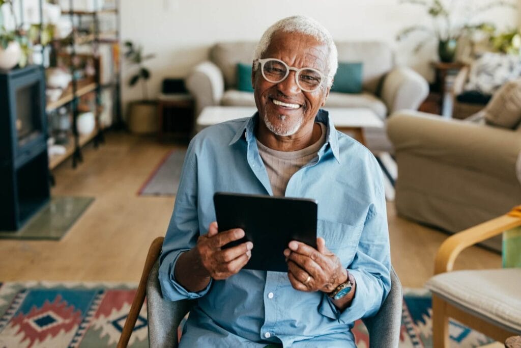 An older adult man sits in a chair, holding a tablet and smiling at the camera.