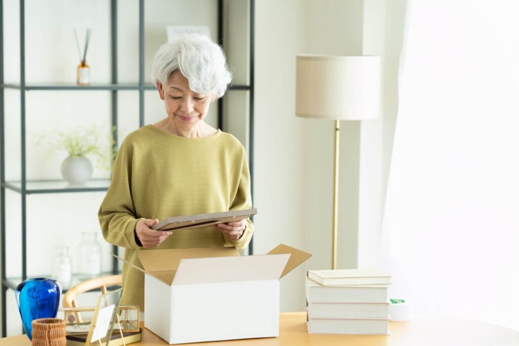 An older adult woman stands at a table with various items on in. She is packing a picture frame into a box.