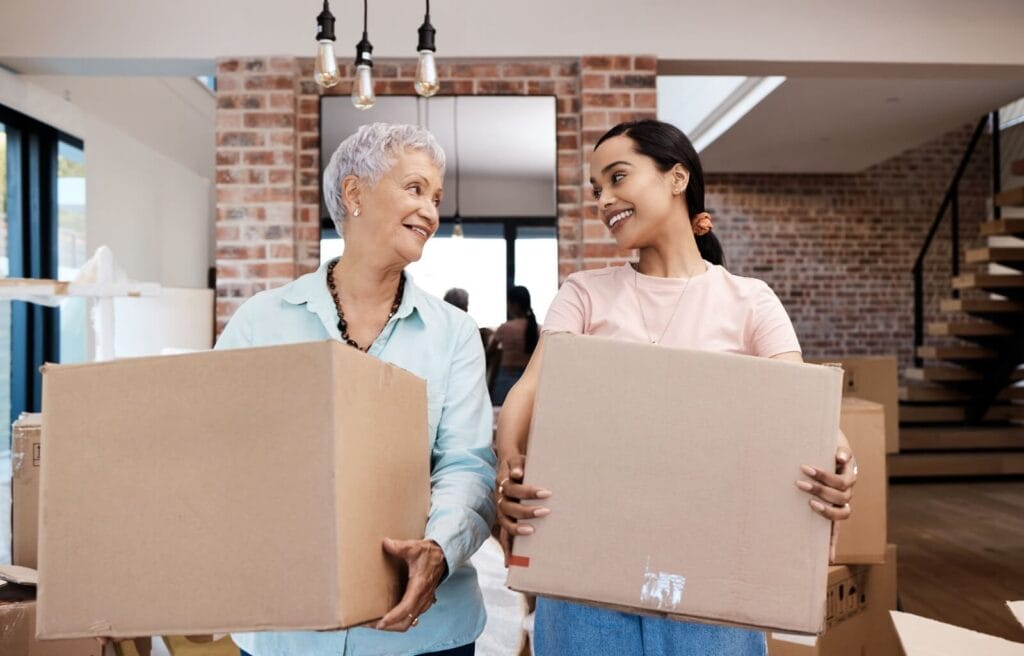 A woman and her older adult mother are standing inside a home, holding cardboard boxes and smiling at each other.