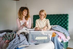 A woman and her older adult mother are sitting on a bed, sorting through clothing. There is a bin labeled "DONATION" in front of them.