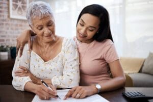 A woman and her older adult mother are sitting at a table. The younger woman has her arm around the older woman's shoulders. They are working on paperwork together. There is also a calculator on the table.