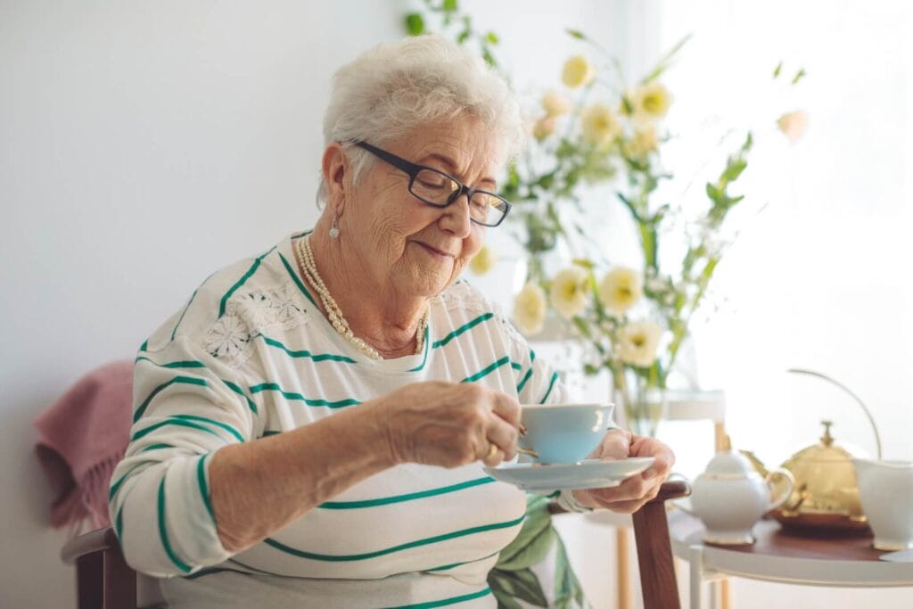 An older adult woman sits in her senior living apartment, drinking a cup of tea. On a table beside her are flowers, a teapot, a sugar dish, and a pitcher for cream.