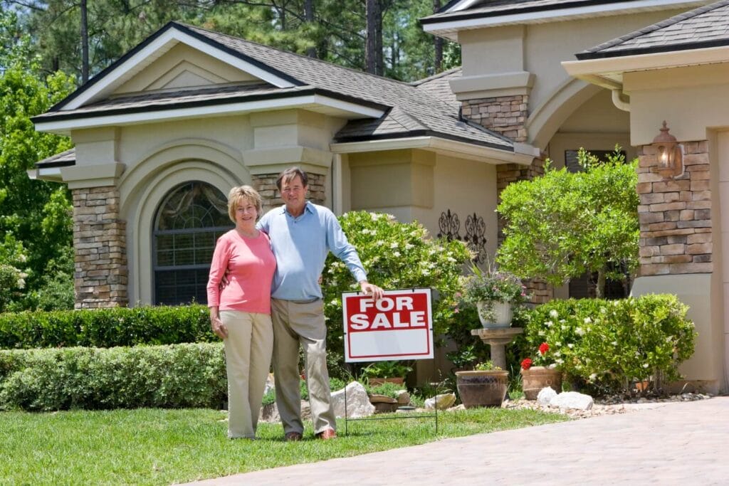 An older adult man and woman stand in front of their house, which has a "For Sale" sign in the yard.