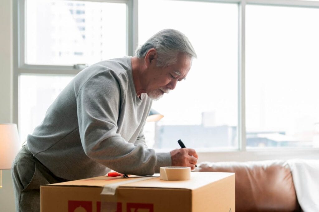 An older adult man is writing on a cardboard box he has packed for a move.