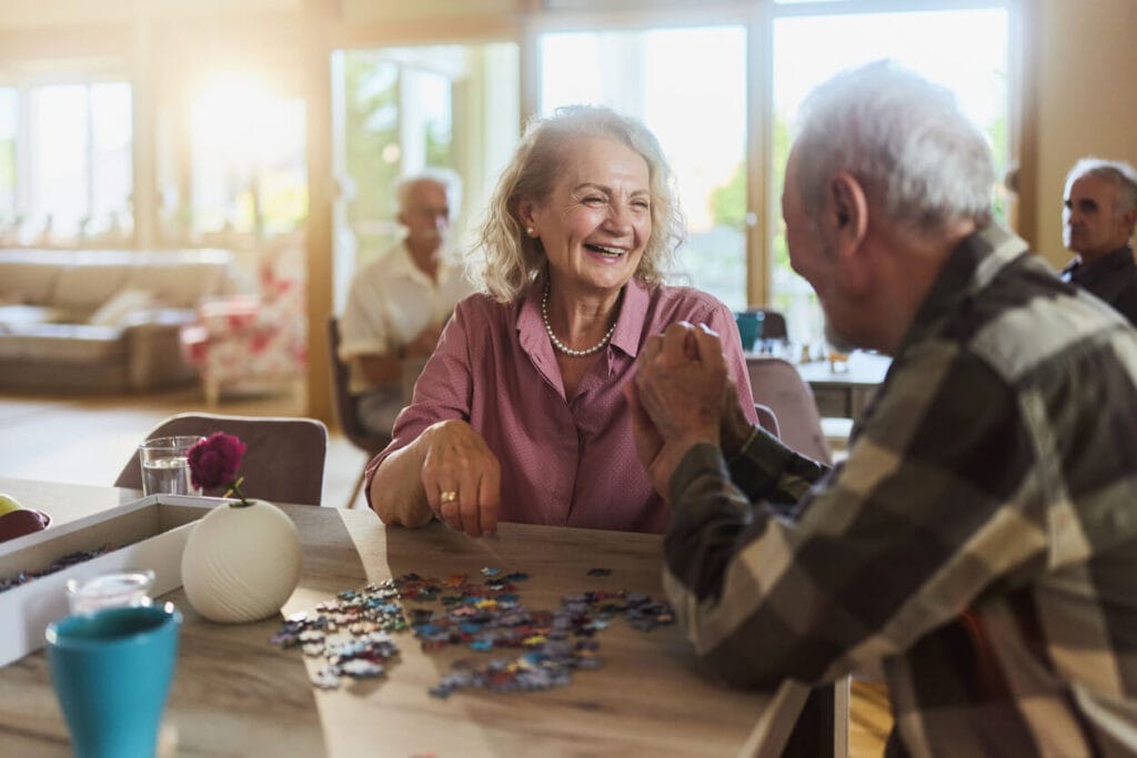 A woman and man sit at a table with jigsaw puzzle pieces on the table. The woman smiles happily at the man, and he appears to smile back.