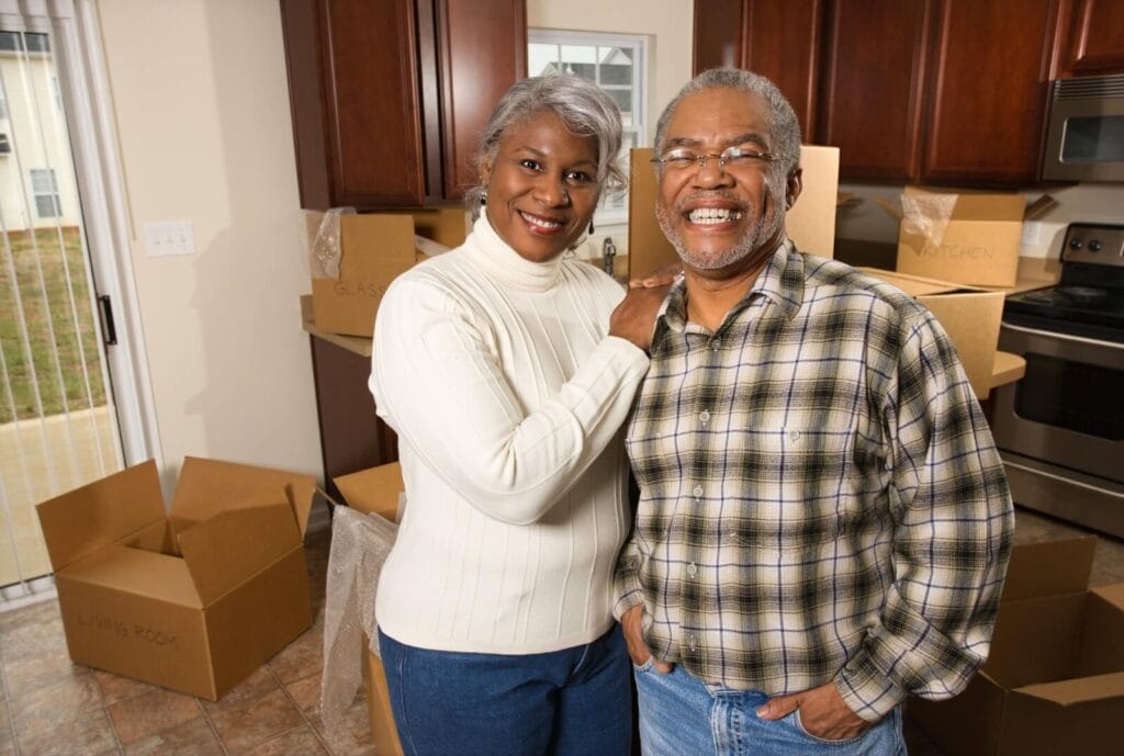 An older adult man and woman stand in a kitchen with carboard boxes scattered around them on the floor and counters. They are smiling at the camera.