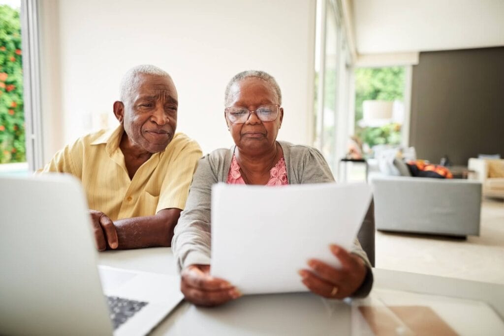 An older adult man and woman sit at a table with a laptop on it, looking at some paperwork.